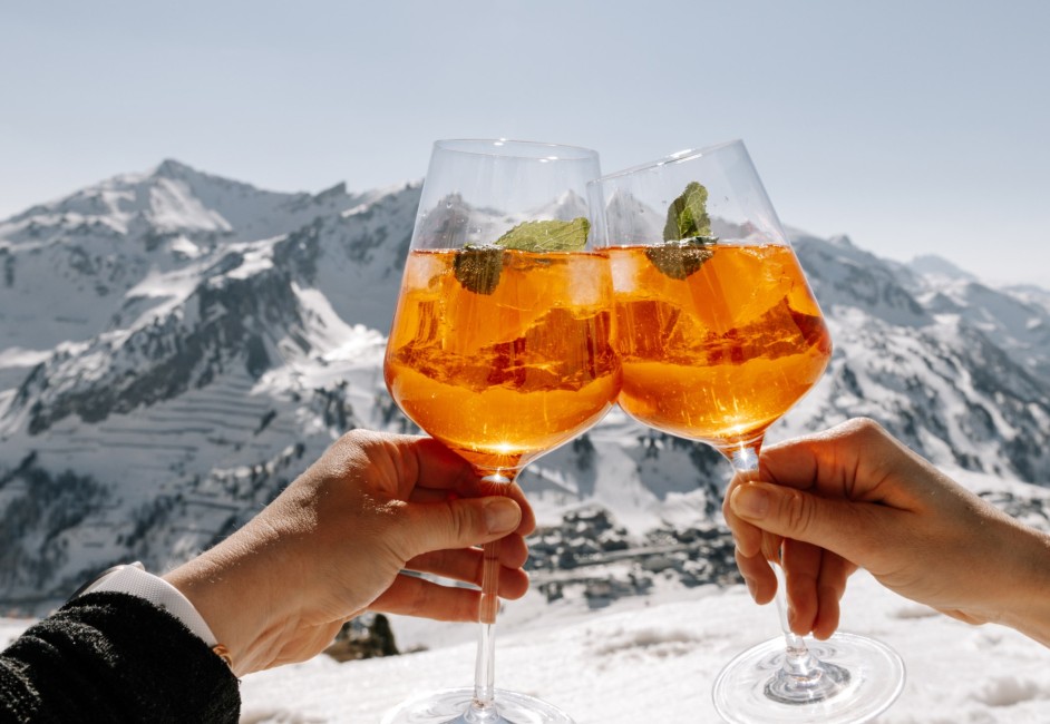 Zwei Personen stoßen mit Aperol Spritz vor verschneiten Bergen in Obertauern an ©TVB_Obertauern