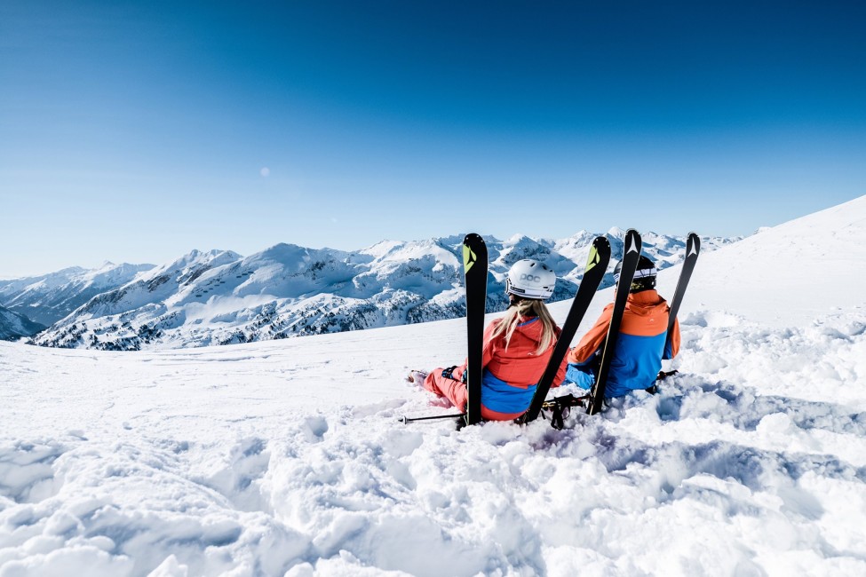 Zwei Personen sitzen mit Skiern im Schnee, blicken auf eine verschneite Berglandschaft Obertauerns ©TVB_Obertauern