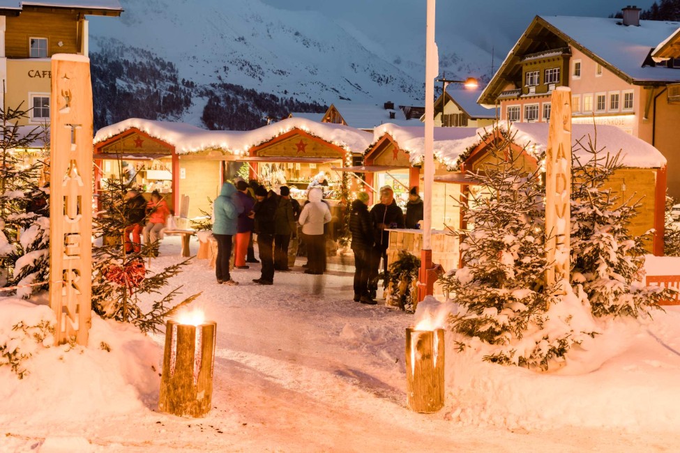 Ein Weihnachtsmarkt in verschneiter Alpenkulisse mit beleuchteten Ständen und Besuchern in Obertauern. ©TVB_Obertauern