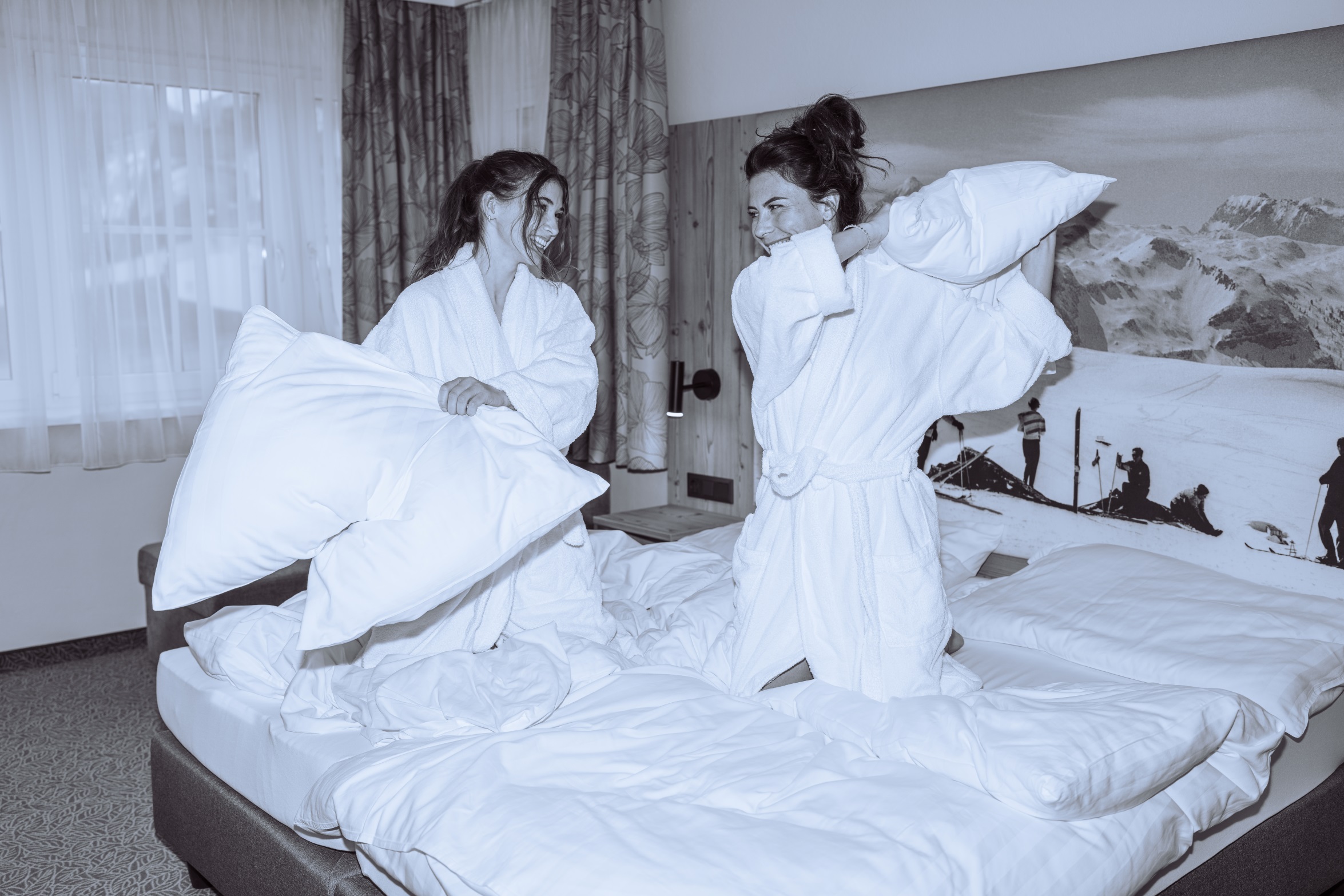 Schwarzweißbild einer Kissenschlacht zwei junger Frauen im Bademantel im Schlafzimmer des Wagner Hotels in Obertauern.