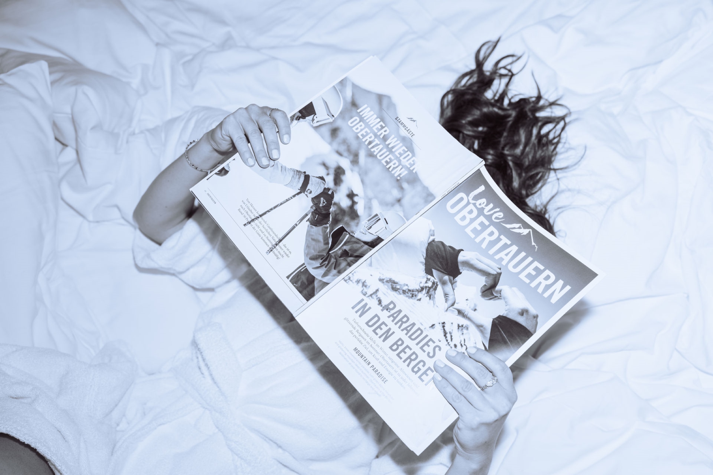 Schwarzweißbild in der Vogelperspektive, ein Gast entspannt im Bett der Wagner Hotels in Obertauern und liest ein Magazin nach einem Tag im Schnee.