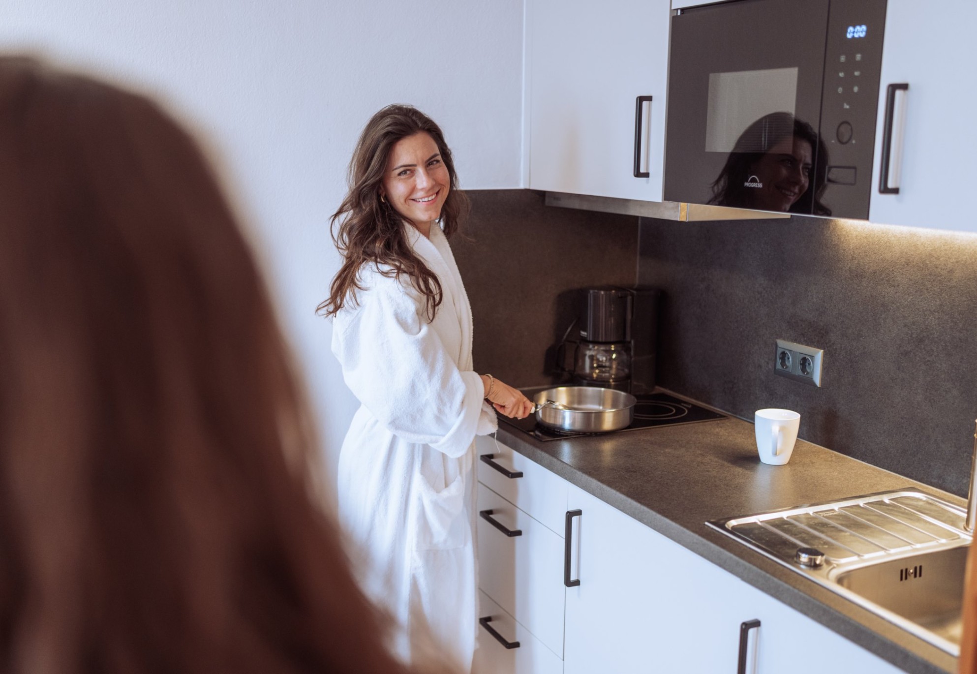 Eine lächelnde Frau in einem Bademantel steht in der Küche und kocht