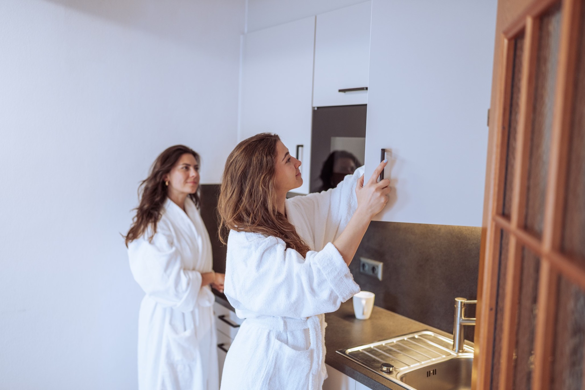 Zwei Frauen im Bademantel in einer Küche, eine öffnet einen Schrank