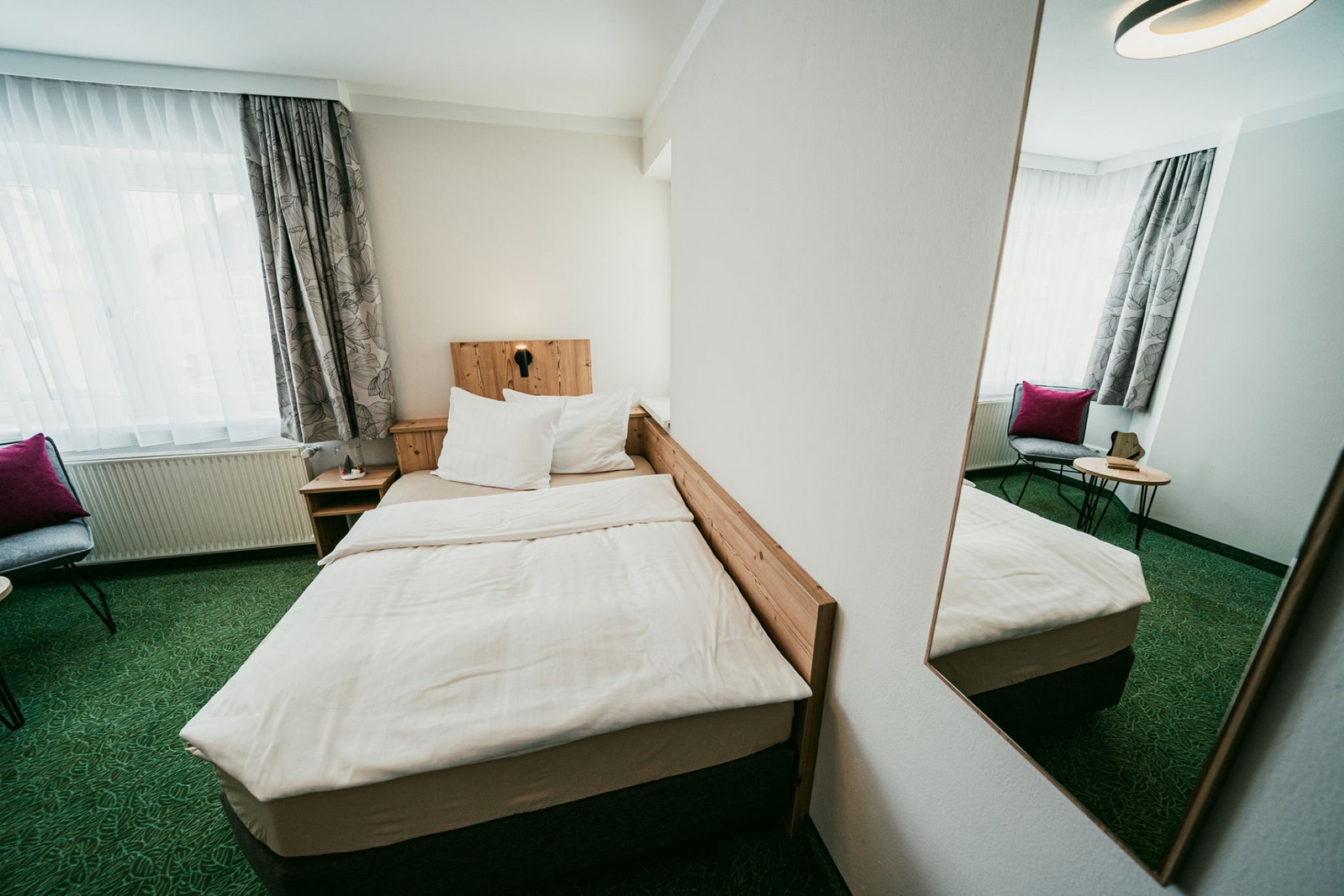 Helles Hotelzimmer mit einem Einzelbett, Spiegel, grünem Teppich und Sitzbereich
