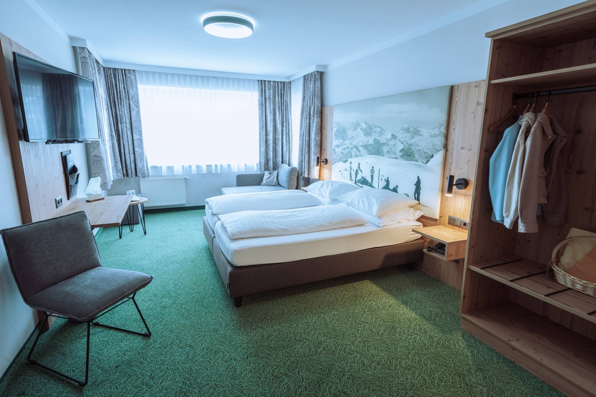 Helles Hotelzimmer mit Doppelbett, Fernseher, Sitzgelegenheit und offener Garderobe