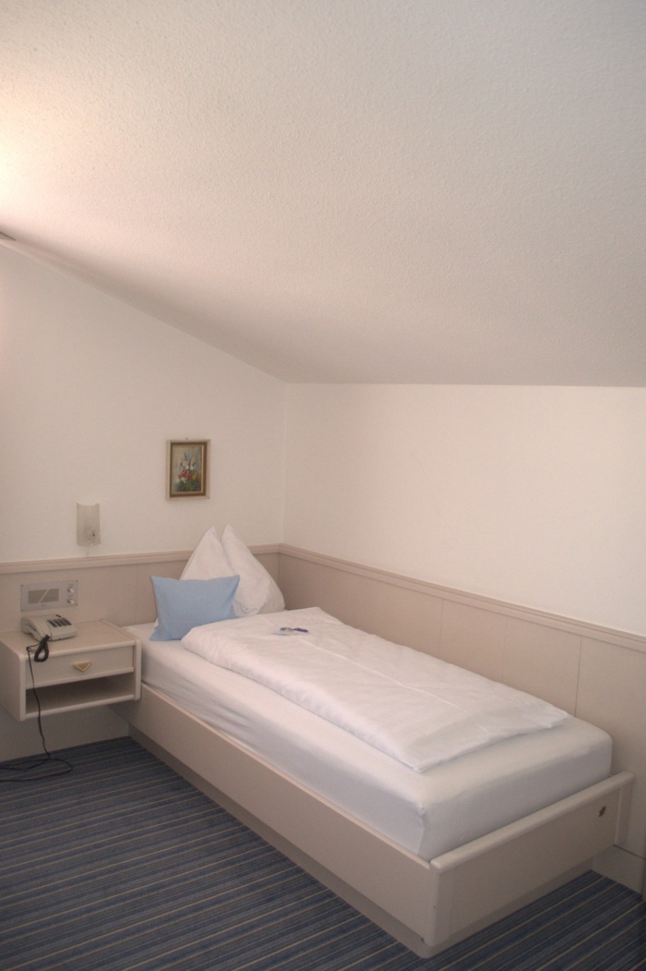 Ein schlichtes Einzelzimmer mit einem schmalen Bett und einem kleinen Nachttisch