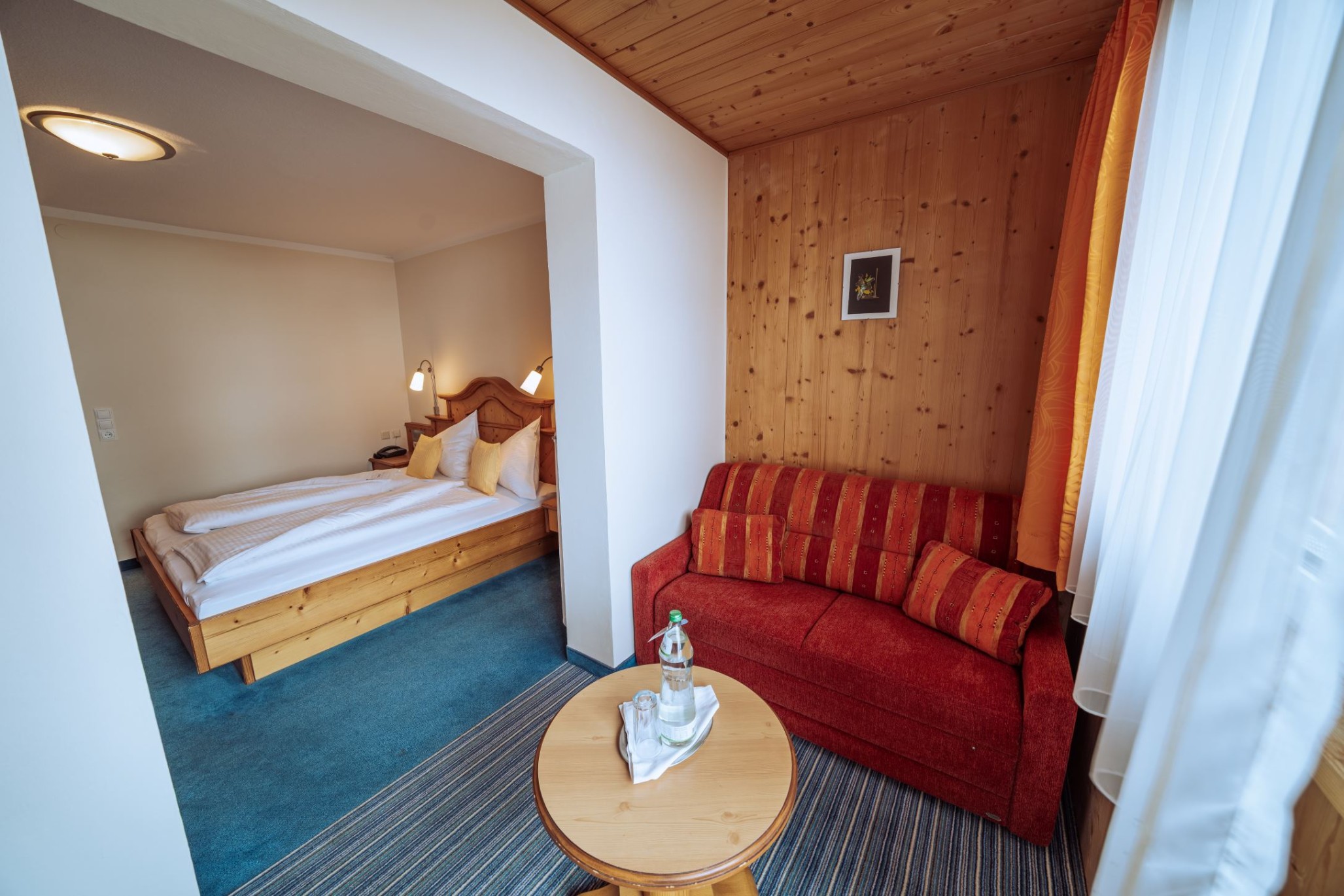 Hotelzimmer mit Doppelbett, rotem Sofa und Tisch mit Wasserflasche