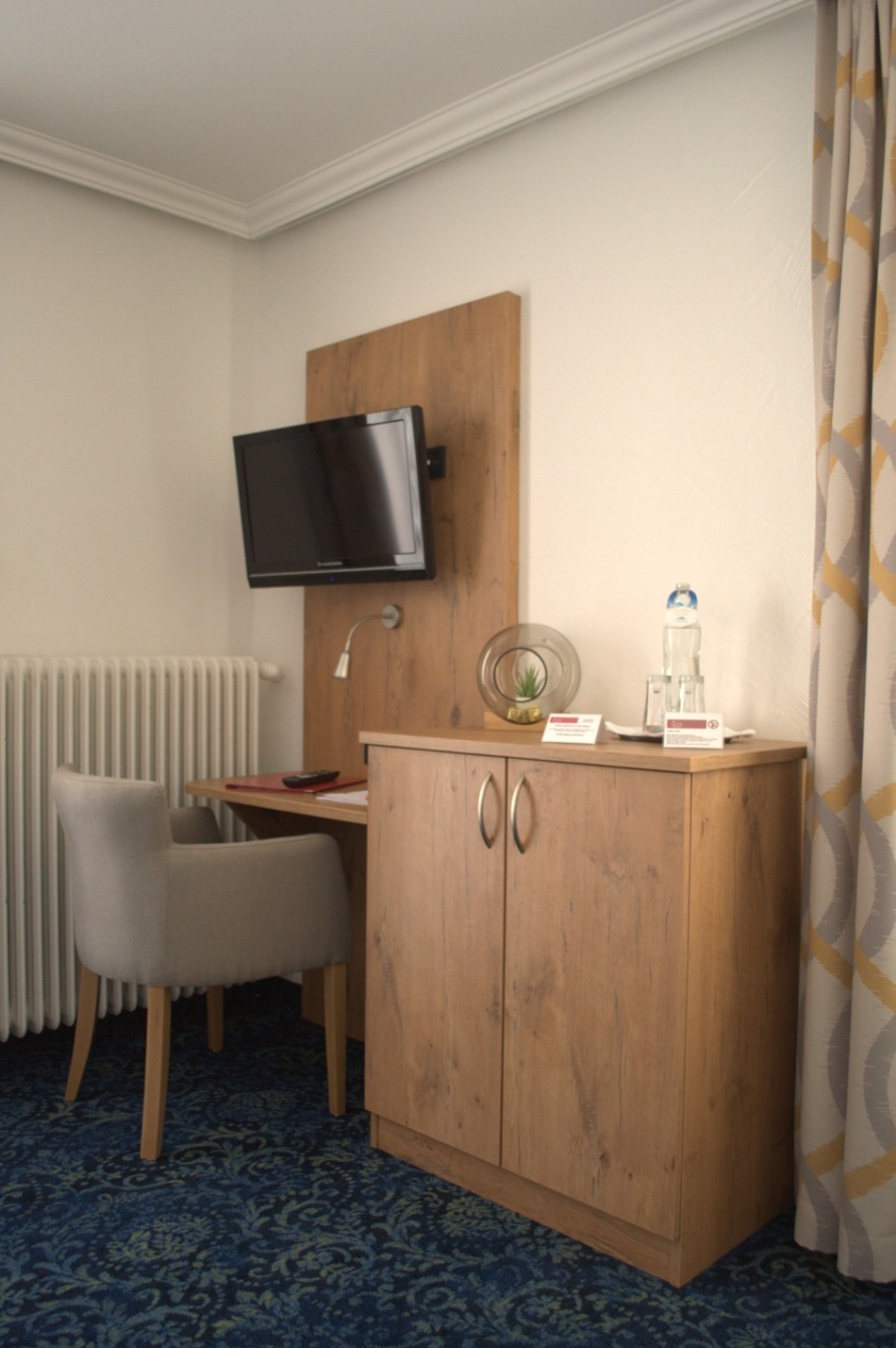 Ein schlichtes Hotelzimmer mit Schreibtisch, TV und Wasserglas auf einem Schrank